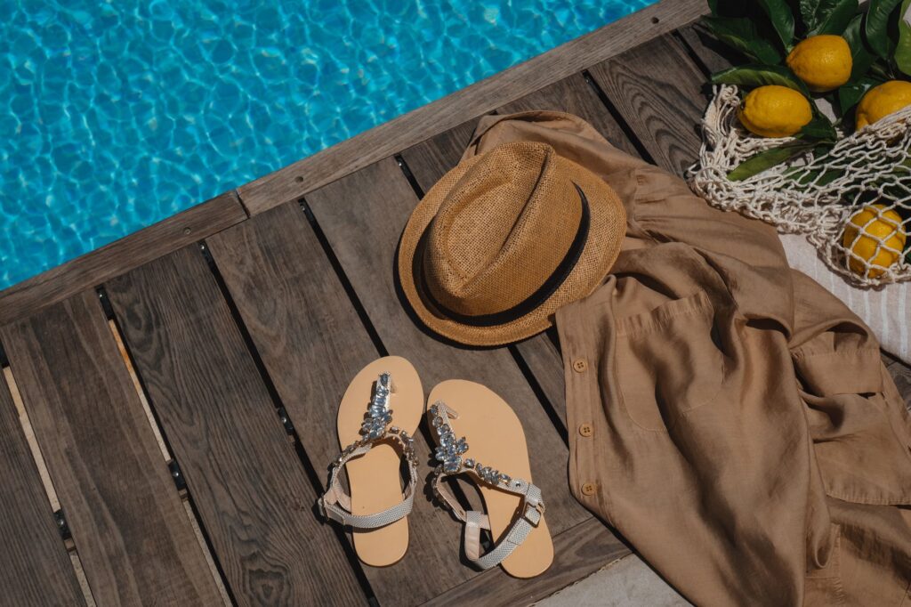 Sandales décorées, chapeau et chemise en lin au bord d'une piscine ensoleillée.
